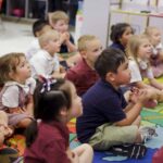 a number of young Kindergarten students in a classroom sit on the floor while focused on something out of frame