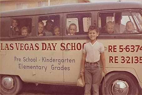 a small bus labeled "Las Vegas Day School" filled with kids looking out of the windows while a young student stands outside of it