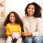 mother and daughter holding piggybank sitting on couch