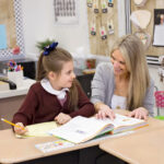 a smiling teacher seated at a desk with a student has her hand on a workbook