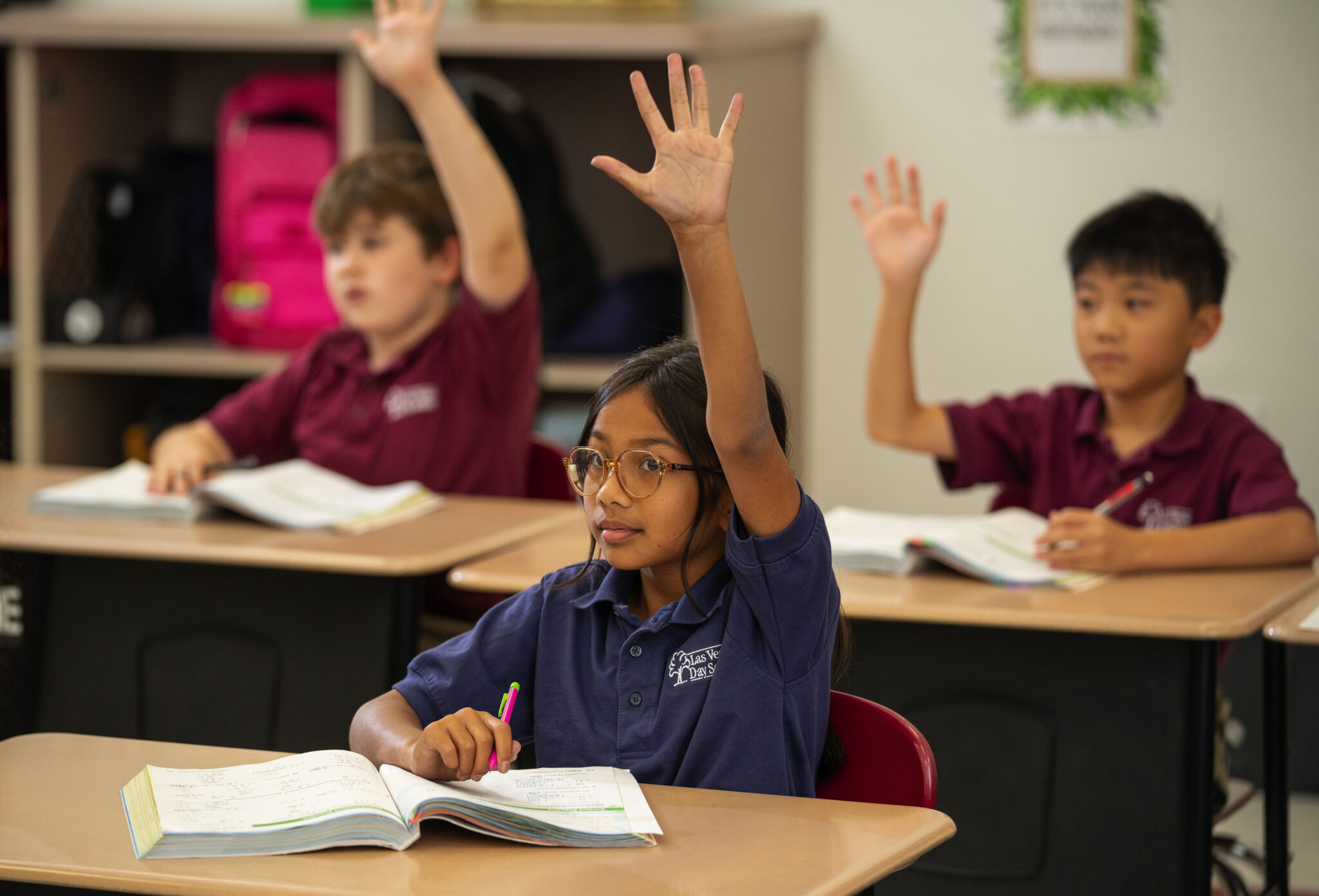 3 young students raise their hands at their desks at Las Vegas Day School