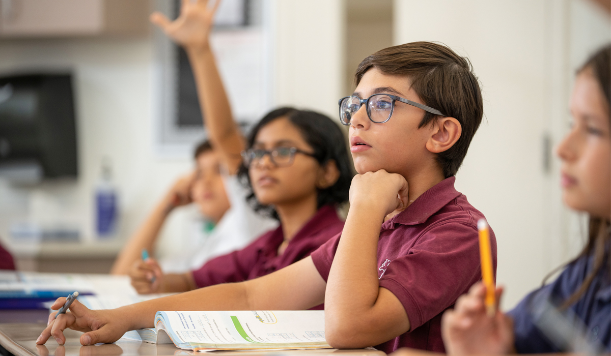 a young boy with glasses at a school desk looks forward as another student in the background raises their hand