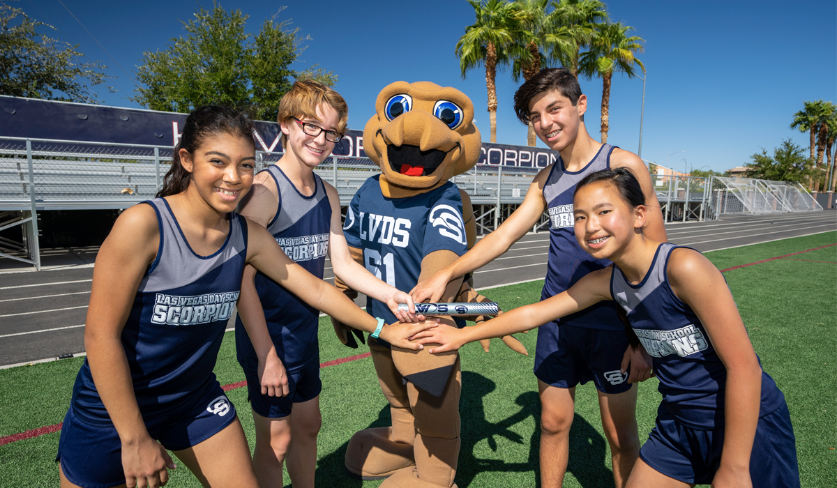 4 young student athletes in blue track uniforms along with the school mascot scorpion putting their hands in to touch a track baton as the smile for the camera