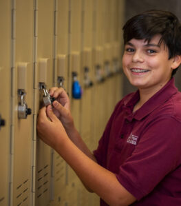 a young student with braces smiles at the camera while he unlocks the padlock on his locker