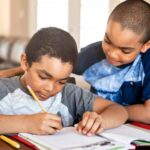 a young student looks over the shoulder of another student who is writing in a workbook