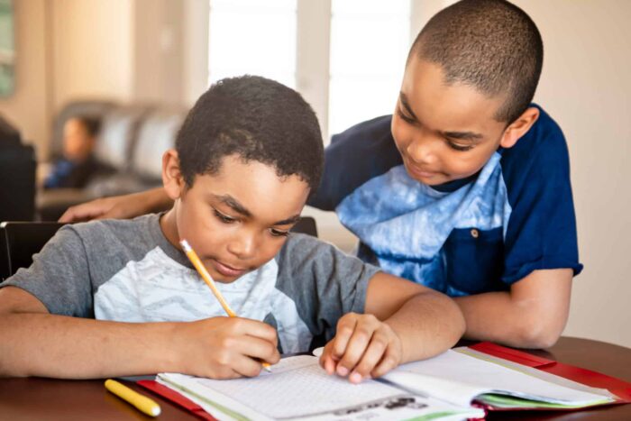 a young student looks over the shoulder of another student who is writing in a workbook