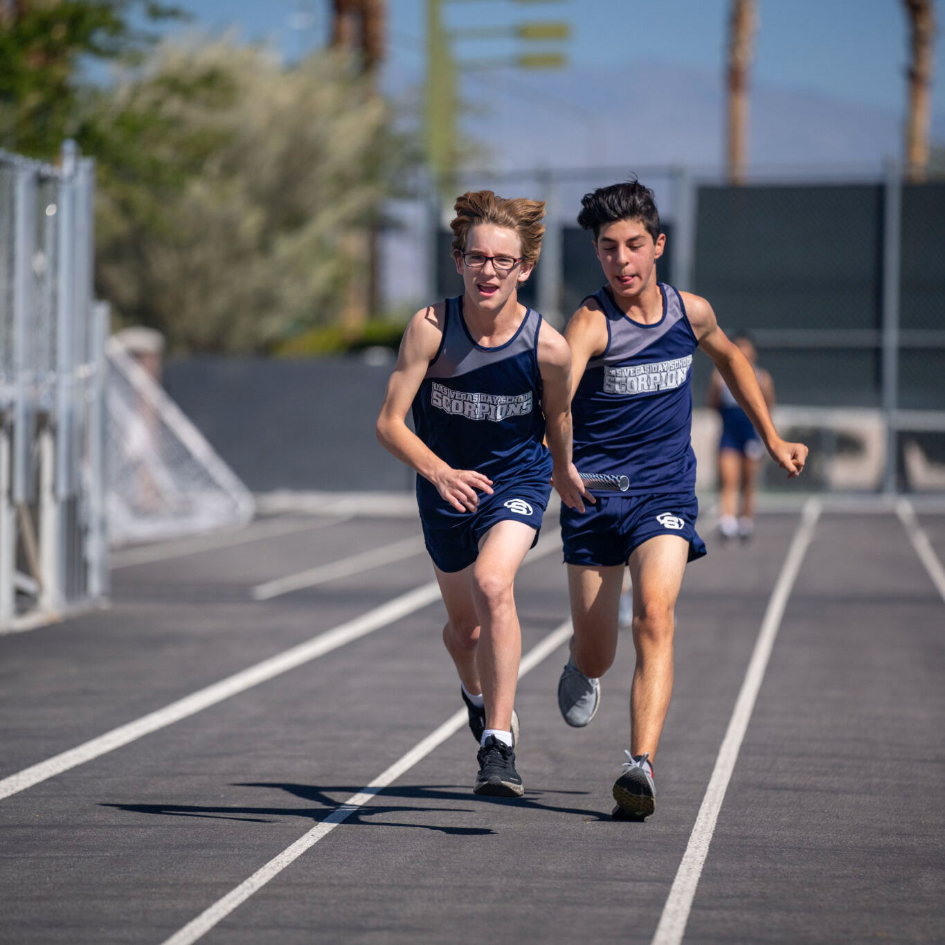 a student in a blue track uniform passing a baton to another track runner during a relay race at a school track